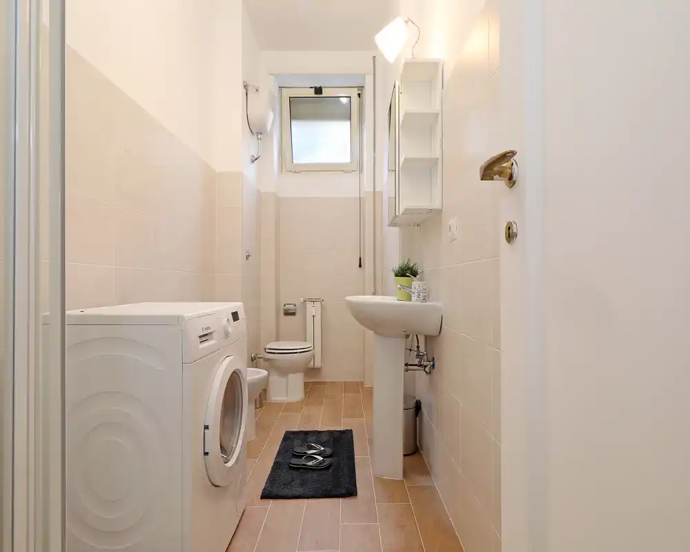 Well-lit modern bathroom with a pedestal sink, toilet, washing machine and small window — clean neutral tiles and wood-effect floor create a fresh, functional space.