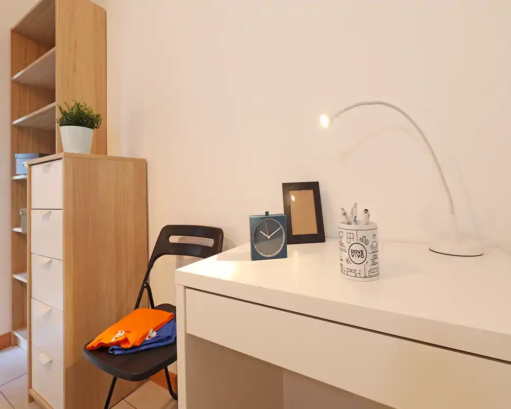 Angolo studio luminoso e minimal in camera da letto con scrivania bianca, lampada regolabile, sveglia e scaffalatura ordinata — un piccolo spazio di lavoro compatto.