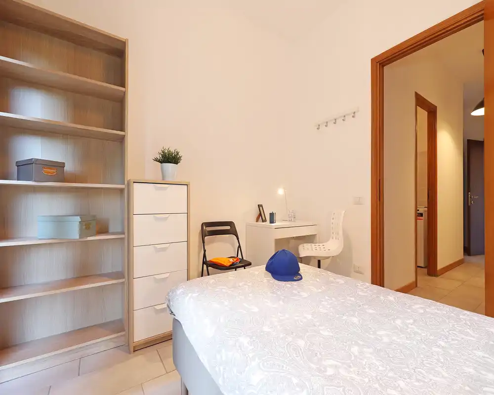 Camera da letto luminosa e ordinata con letto in primo piano, un'alta libreria a giorno e un comò, oltre a una piccola scrivania e sedie—ideale per soggiorni confortevoli.