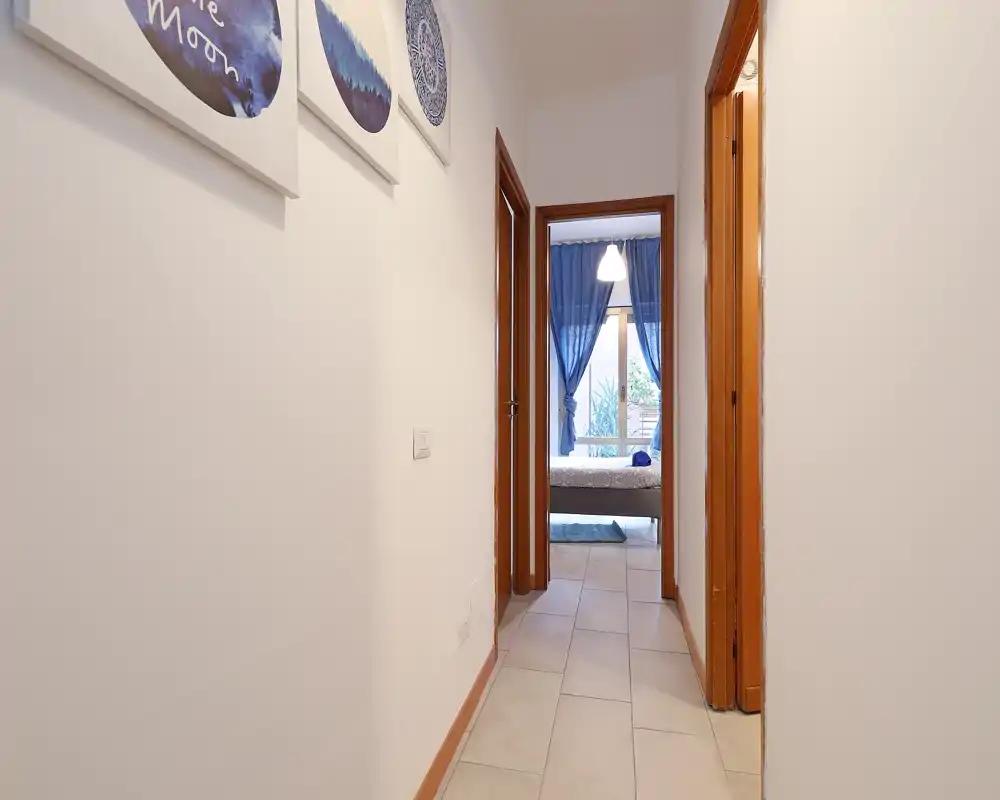 Corridoio che conduce a una camera da letto; si vedono tende blu attraverso la porta. La foto mette in risalto il corridoio e l’ingresso alla zona notte con luce naturale dalla finestra.