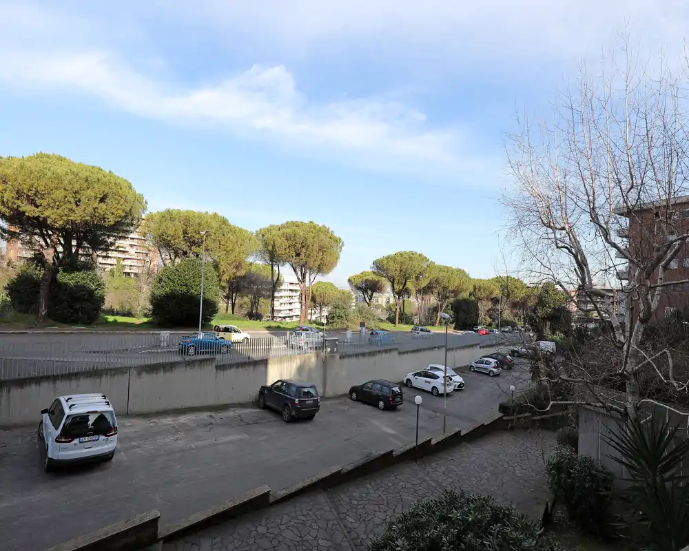Vista esterna di un parcheggio residenziale e di un'area verde adiacente con pini a ombrello sotto un cielo luminoso.