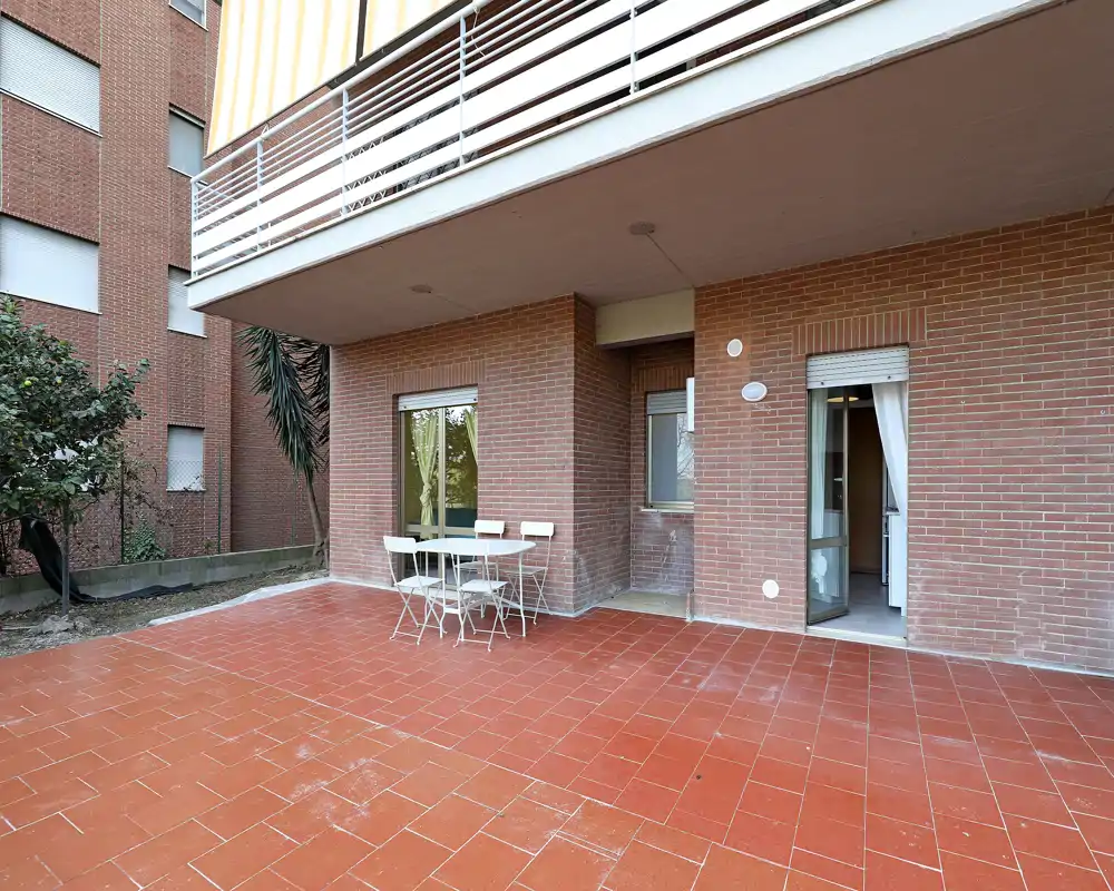 Ampia terrazza a piano terra con pavimento in piastrelle rosse, piccolo tavolo e sedie da esterno e accesso diretto all'appartamento tramite porta scorrevole.