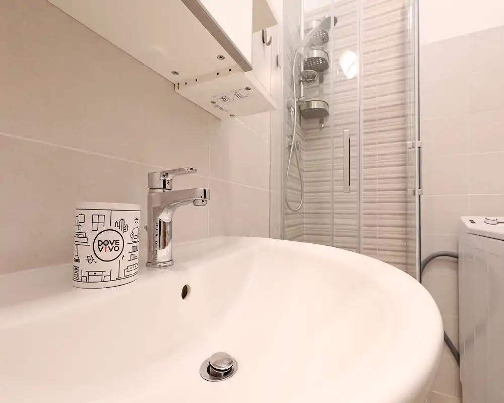 Bagno moderno e ben illuminato con primo piano del lavabo bianco, rubinetto cromato, box doccia in vetro e la lavanderia visibile sul bordo.