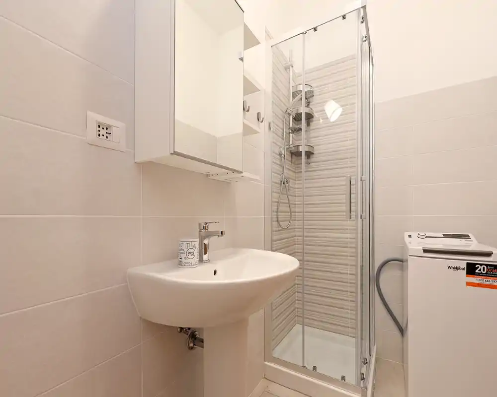 Bagno compatto e pulito con lavabo a colonna, mobiletto specchiato e box doccia angolare in vetro; piastrelle beige neutre e lavatrice a carica dall'alto completano la disposizione funzionale.