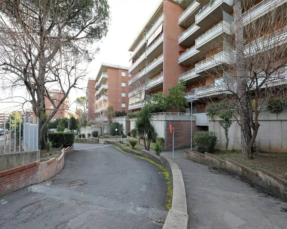 Veduta esterna dell'ingresso e della corsia di un complesso residenziale con palazzine in mattoni, balconi, strada asfaltata e aiuole.