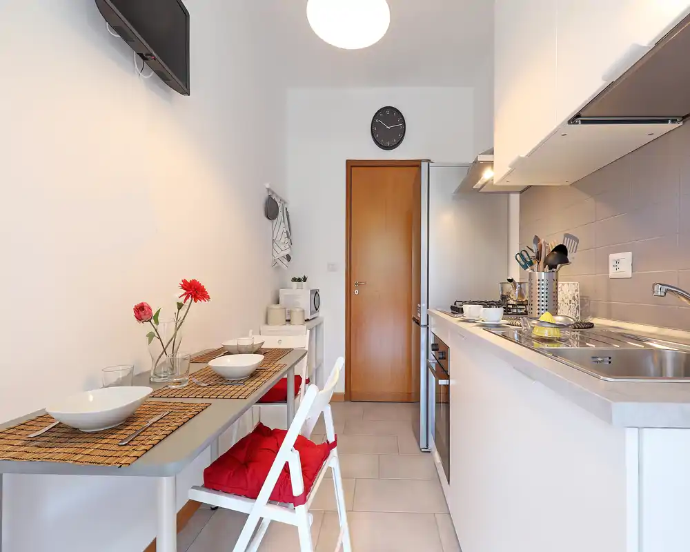 Cucina moderna compatta con piano lineare e piccolo angolo pranzo. Si vedono lavello in acciaio, piano cottura, frigorifero e un tavolo per due apparecchiato con tovagliette e fiori, perfetta per un annuncio di appartamento.