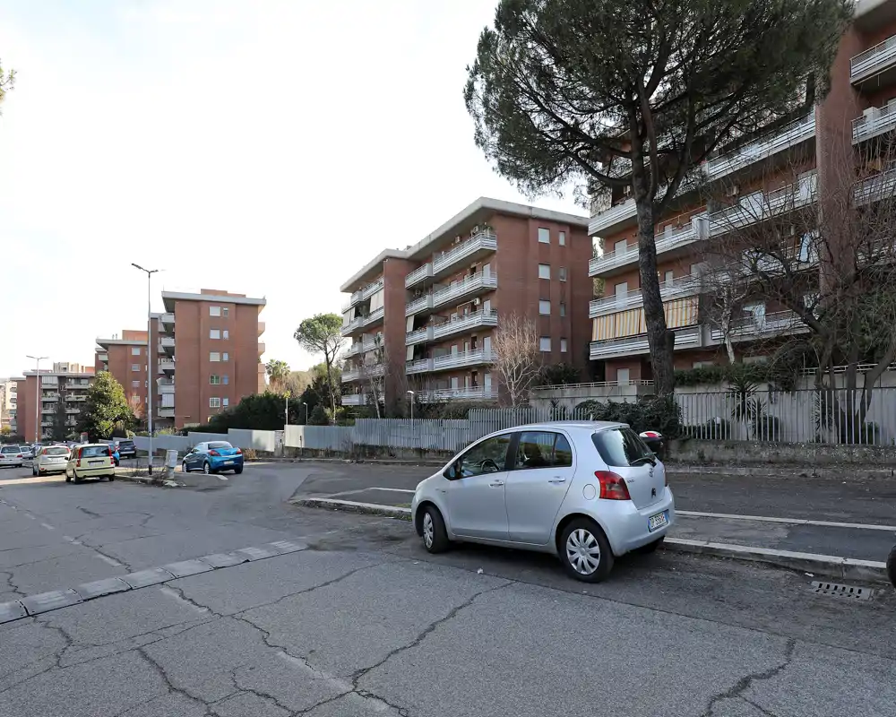 Vista stradale di edifici residenziali con auto parcheggiate e marciapiede ampio; luce diurna intensa e contesto urbano nitido.