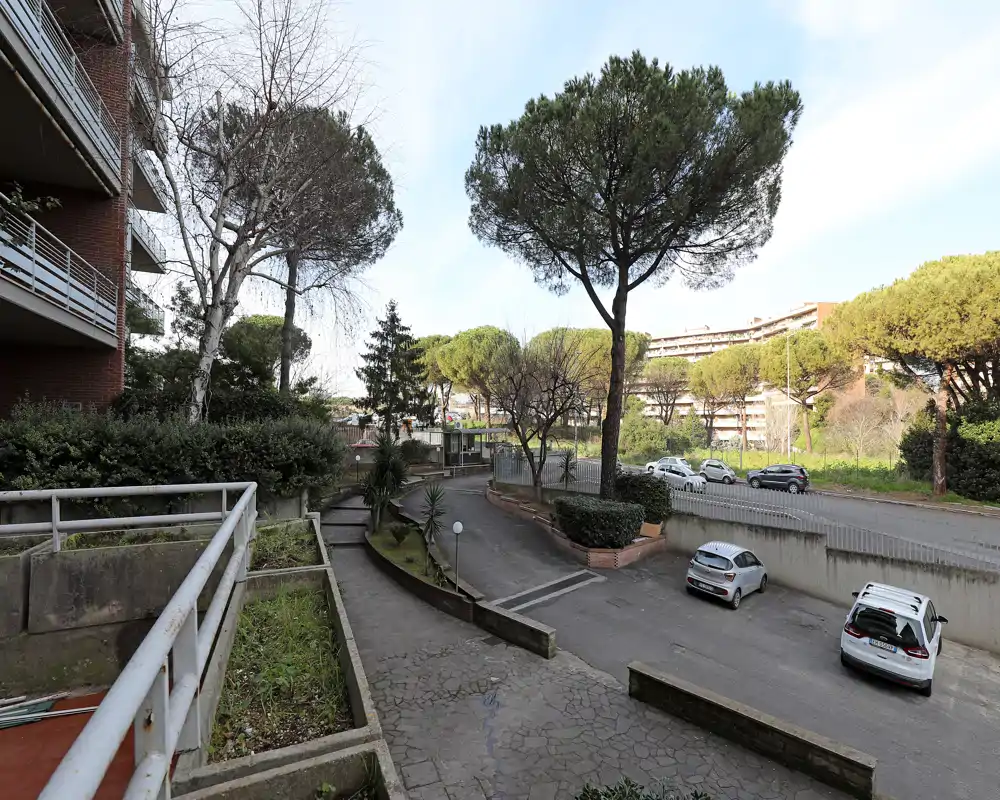 Vista esterna dell'ingresso residenziale e dell'area parcheggio con alberi, fioriere e auto parcheggiate. Giorno luminoso e pavimentazione ben visibile verso la strada recintata.
