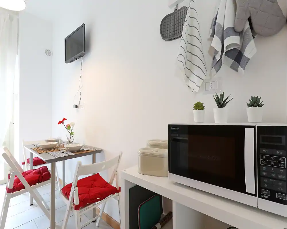 Angolo cottura compatto e luminoso con microonde su scaffale bianco, piccolo tavolo da pranzo per due e cuscini rossi: stile moderno e pulito, ideale per soggiorni brevi.