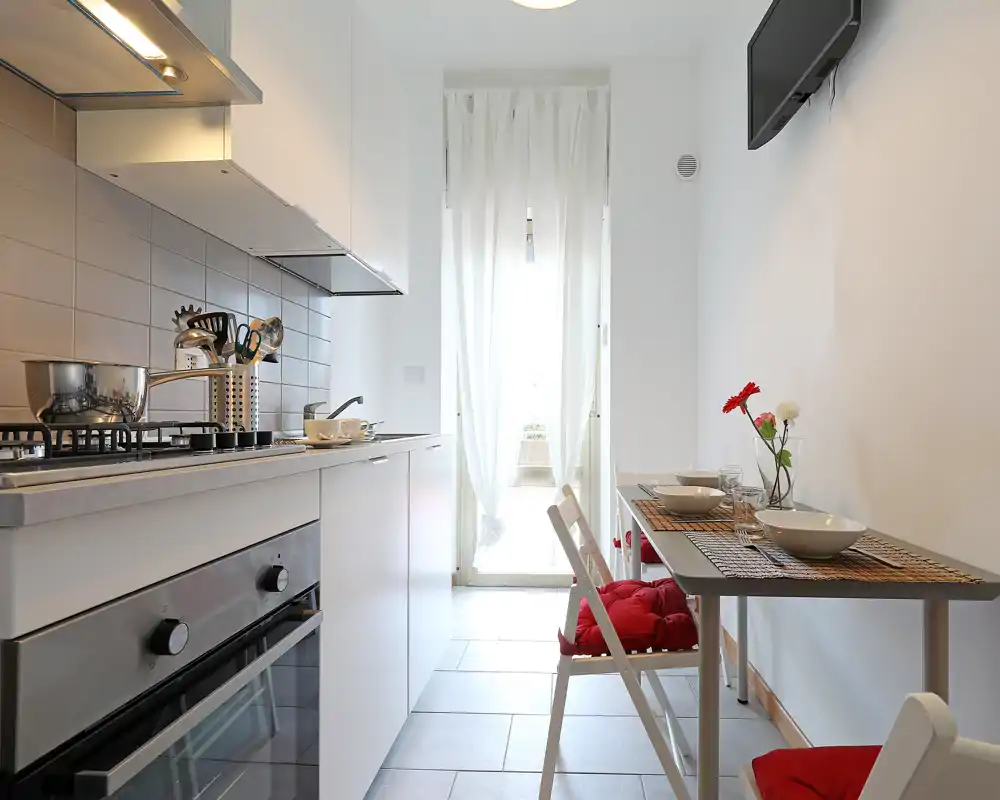 Cucina moderna compatta con pensili bianchi, piano cottura a gas e forno incassato; piccolo tavolo da pranzo vicino alla parete con sedie cuscinate rosse e fiori freschi, luce naturale luminosa dalla porta.
