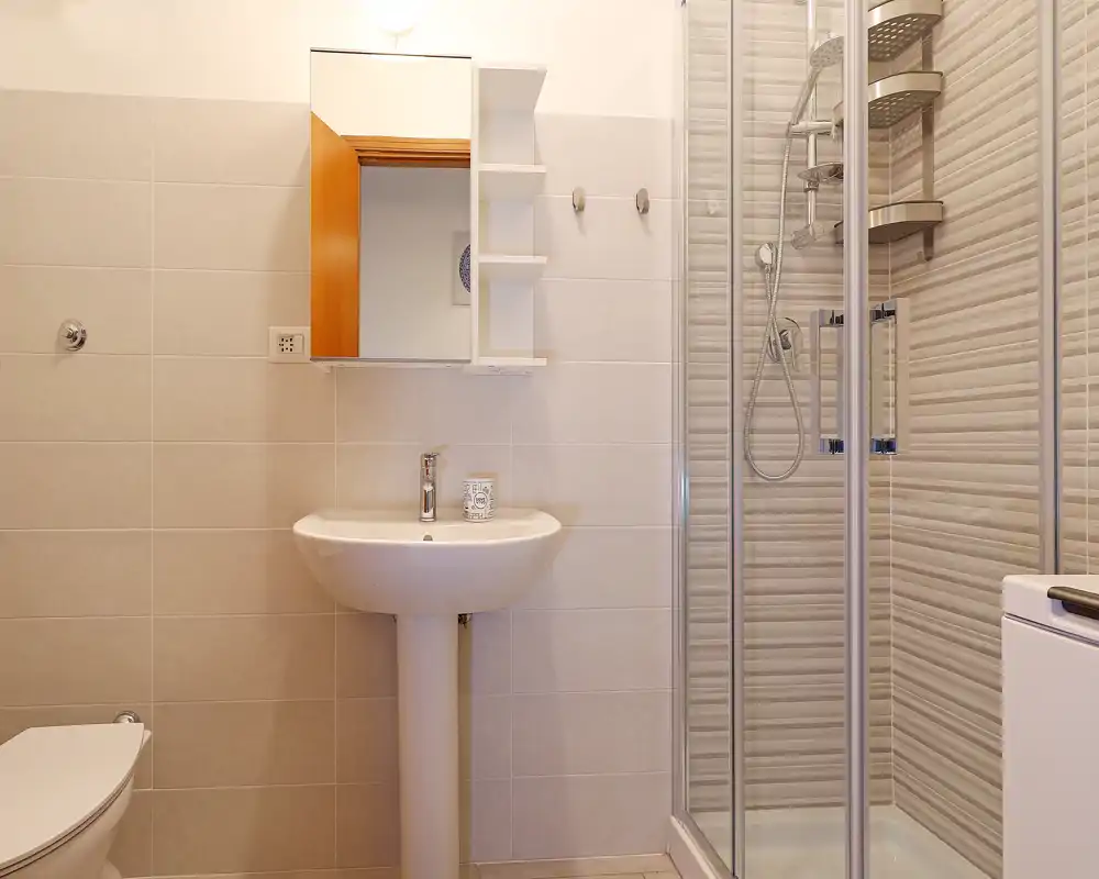 Bagno luminoso e pulito con lavabo a colonna, specchiera con mobiletto e cabina doccia in vetro con rubinetteria moderna.