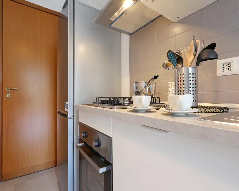 Cucina compatta e moderna con piano cottura a gas, forno incorporato e frigorifero in acciaio. Piastrelle e piano neutri con tazze e utensili in esposizione per un aspetto ordinato e contemporaneo.