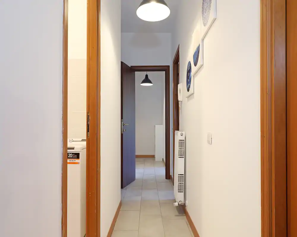 Ingresso/corridoio pulito e stretto con pavimento piastrellato, pareti bianche, telai delle porte in legno e un radiatore sulla parete destra. La visuale arriva in una stanza interna con un lampadario a sospensione.