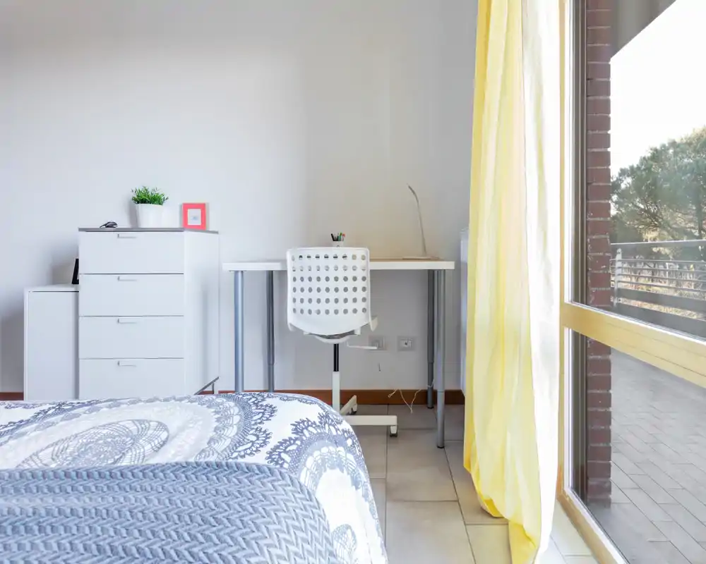 Angolo di camera luminoso e moderno con letto visibile, una piccola postazione con scrivania e sedia e una grande finestra/porta che illumina il balcone.