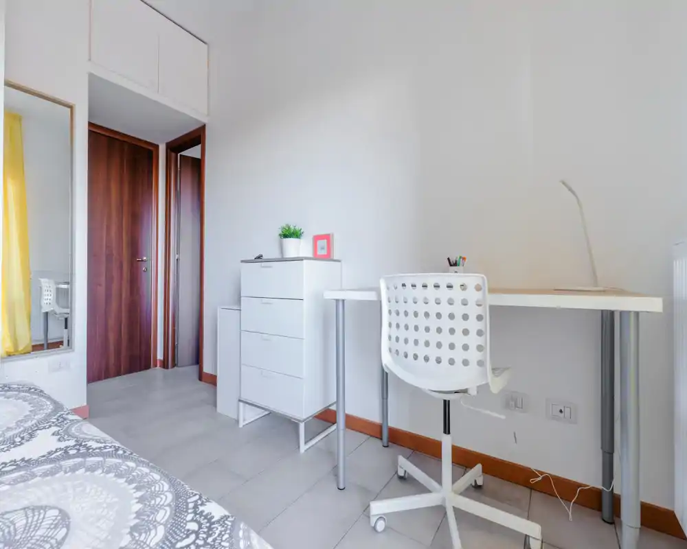 Angolo camera compatto e luminoso con copriletto decorato, cassettiera bianca e una scrivania semplice con sedia — ideale per una persona che cerca uno spazio pulito e ben illuminato.