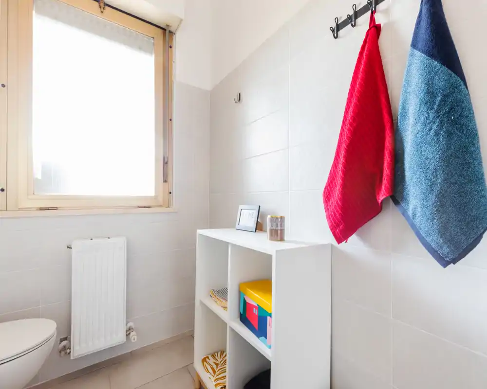Bagno luminoso e pulito con wc, termosifone e ganci a muro con asciugamani colorati; piccolo mobile a cubi per riporre scatole e accessori.