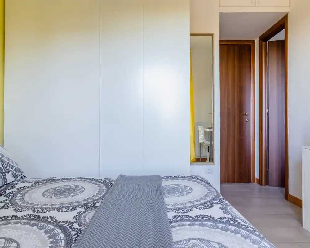 Camera da letto moderna e luminosa con biancheria a motivo e armadio a parete pulito; a destra si vede un corridoio con porte in legno.