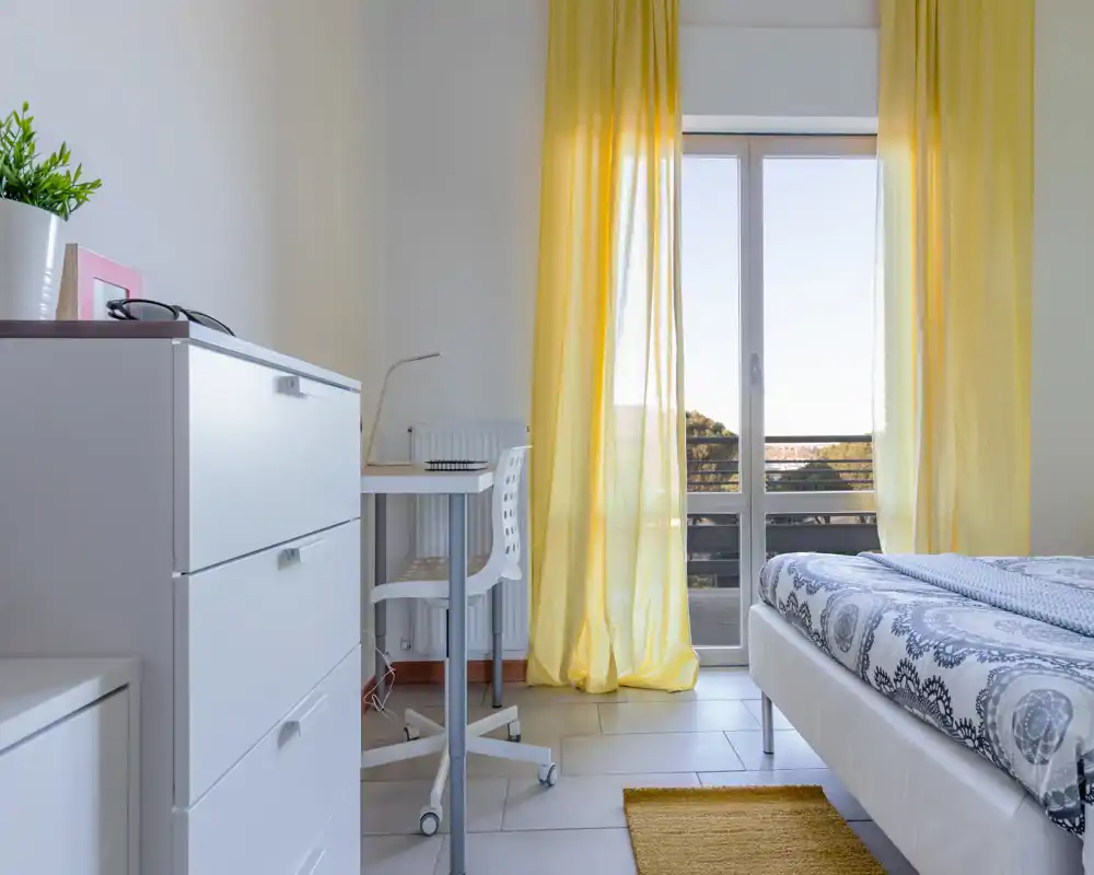 Camera da letto moderna e luminosa con copriletto a motivi, tende gialle e una scrivania accanto alla porta del balcone che lascia entrare molta luce naturale.