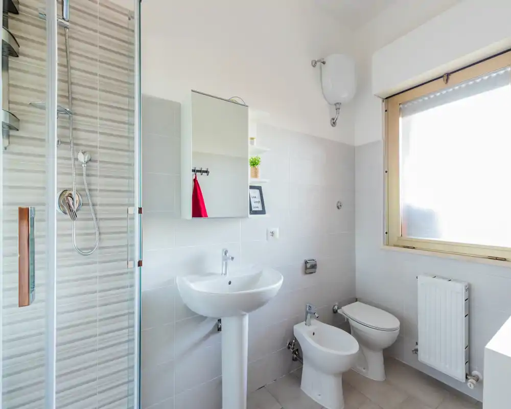 Bagno pulito e luminoso con lavabo a colonna, bidet, WC e doccia con box in vetro. La luce naturale dalla finestra opaca rende l'ambiente arioso e fresco.