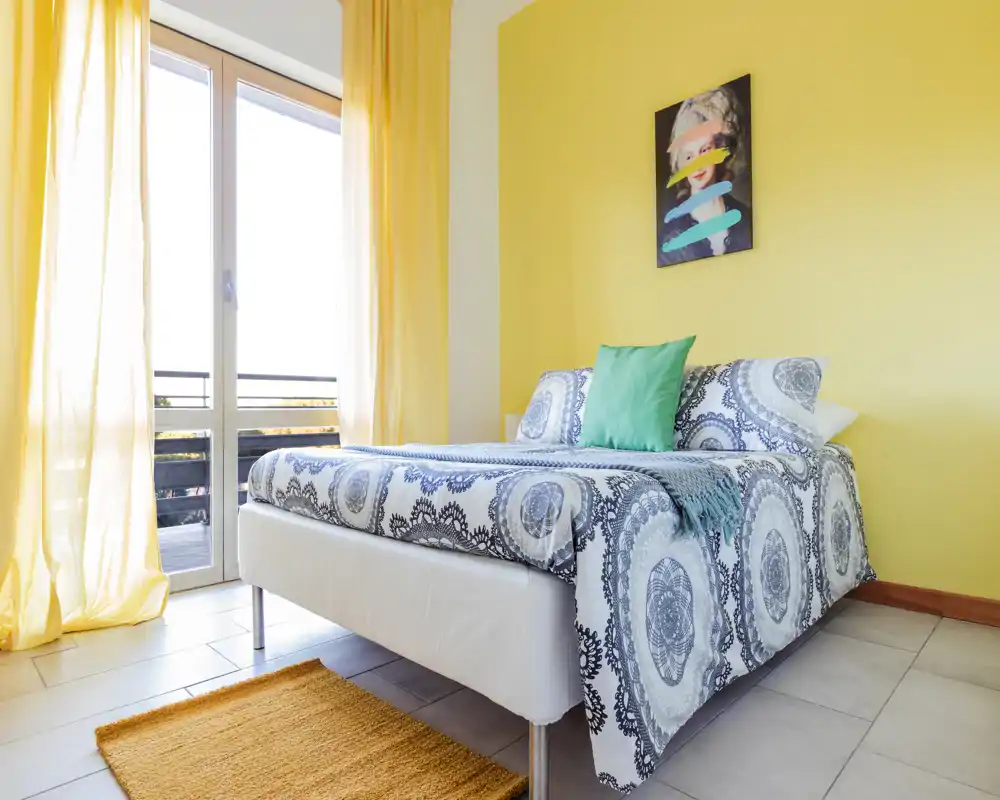 Camera da letto luminosa e moderna con letto matrimoniale dal motivo decorativo, cuscino verde e ampia finestra che illumina la stanza con luce naturale.