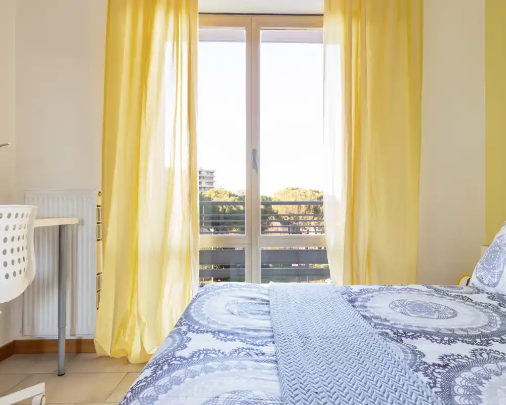 Camera da letto luminosa e ben arredata con copriletto a motivo, plaid morbido e una grande finestra che lascia entrare molta luce naturale.