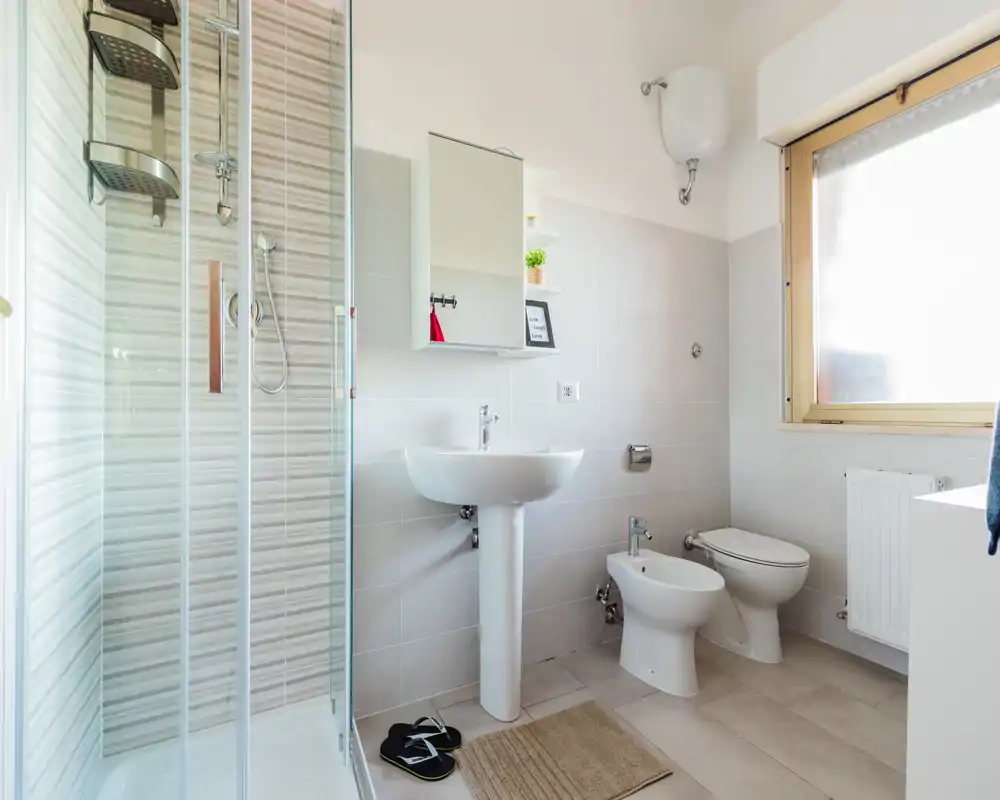 Bagno moderno e luminoso con cabina doccia, lavabo a piedistallo, bidet e WC; piastrelle neutre e una grande finestra satinata garantiscono molta luce naturale.