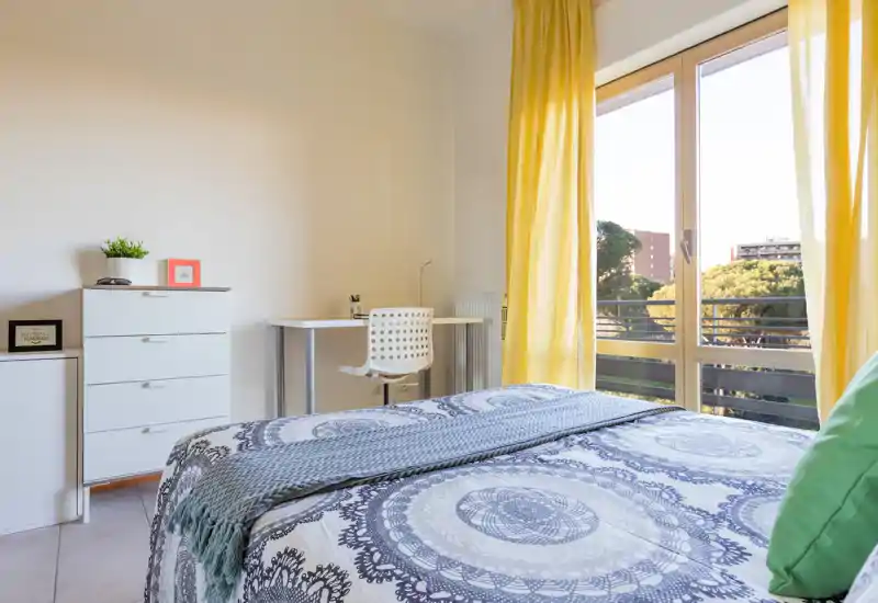 Camera da letto luminosa e ben arredata con un letto matrimoniale dalla fantasia centrale in primo piano, una scrivania e un comò vicino alla finestra e ampie vetrate che lasciano entrare molta luce naturale.