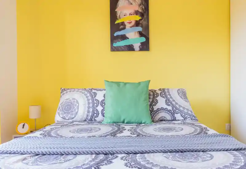 Camera da letto luminosa e di stile con copriletto decorato, cuscino verde centrale e parete gialla vivace; il letto occupa la scena e l'ambiente appare fresco e accogliente.