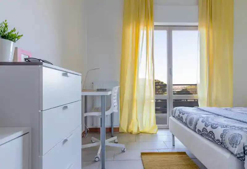 Camera da letto moderna e luminosa con copriletto a motivi, tende gialle e una scrivania accanto alla porta del balcone che lascia entrare molta luce naturale.
