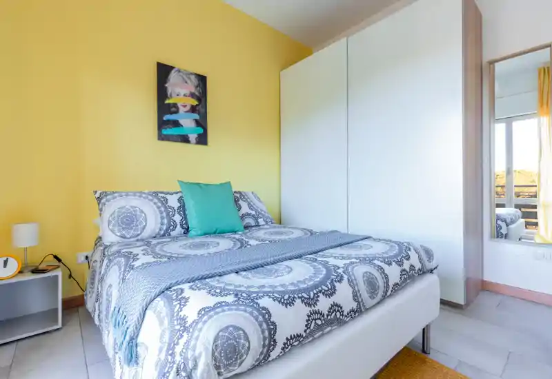 Camera da letto luminosa e moderna con biancheria a motivo, un cuscino turchese e un grande armadio bianco — ben illuminata e arredata con gusto per un soggiorno accogliente.