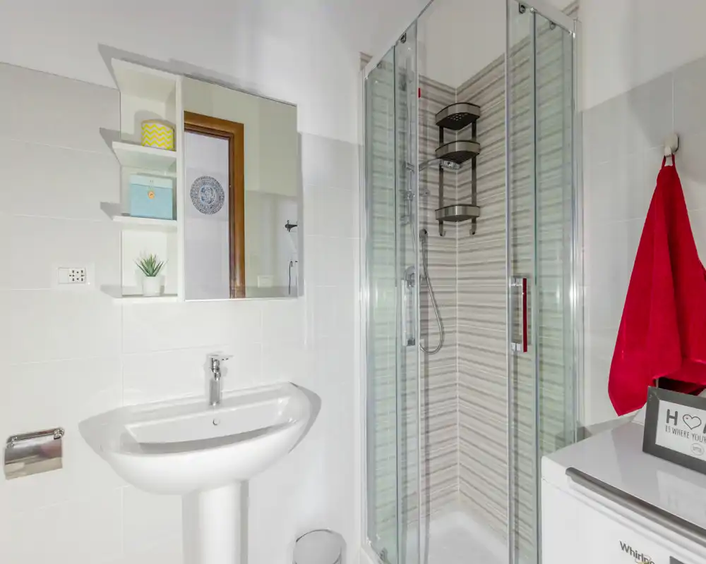 Bagno moderno e compatto con doccia con box in vetro, lavabo a colonna e piastrelle bianche pulite — luminoso e ben tenuto.