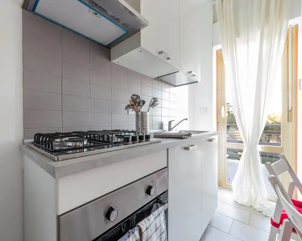 Cucina moderna e luminosa con forno in acciaio, piano cottura a gas e piano compatto; luce naturale da una grande finestra/porta illumina la piastrella e gli utensili.