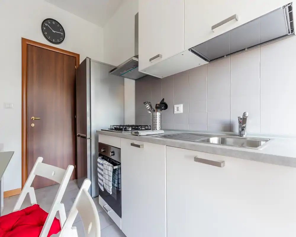 Cucina moderna e luminosa con lavello in acciaio, forno incassato e piano cottura a gas, mobili bianchi e frigorifero — ambiente pulito e funzionale adatto ad affitti brevi.