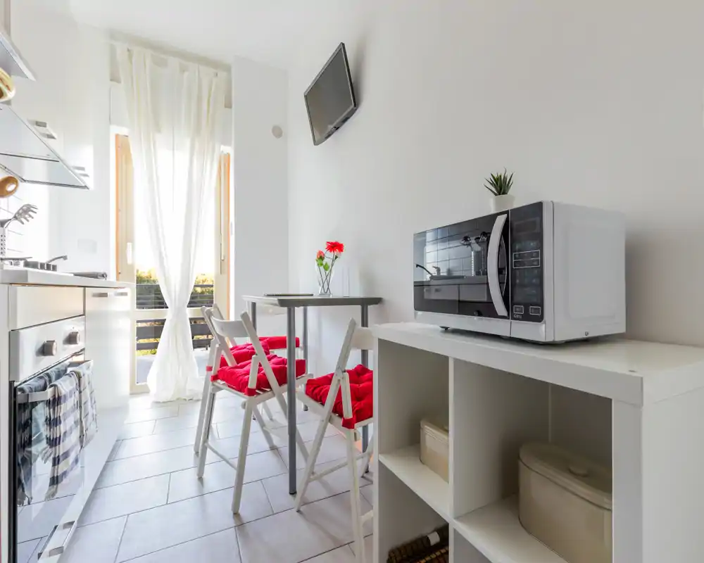 Cucina compatta e moderna con piano cottura, microonde e un piccolo tavolo da pranzo vicino alla porta balcone luminosa; i cuscini rossi e un vaso aggiungono un tocco accogliente.