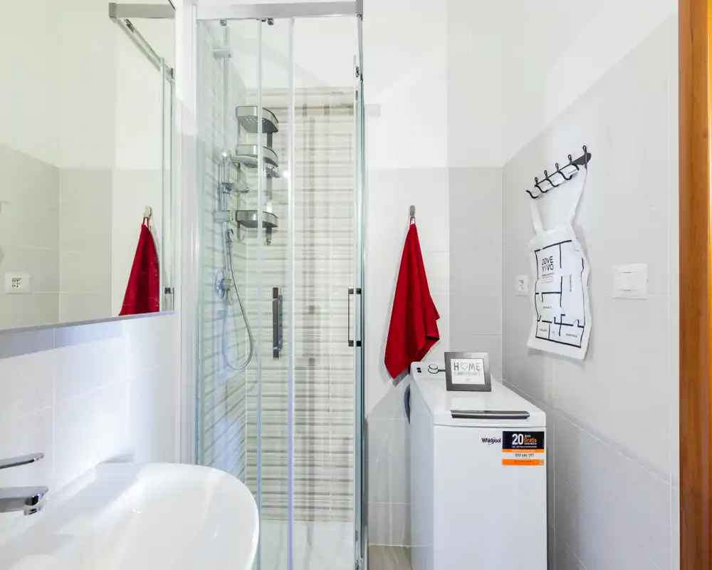 Bagno moderno e compatto con doccia a filo in vetro, lavabo a parete e lavatrice a carica dall'alto; piastrelle neutre luminose e asciugamani rossi per un tocco di colore.