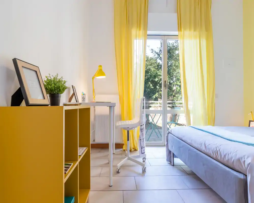 Bright, cozy bedroom with a comfortable bed, a study desk and yellow curtains opening onto a small balcony. Natural light fills the space and warm accents give a welcoming atmosphere.