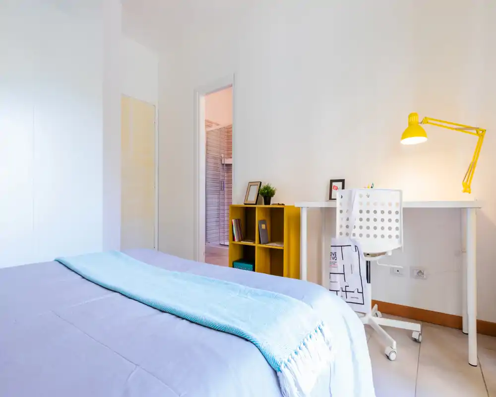 Bright, modern bedroom with a large bed, light-blue throw and a compact workspace featuring a white desk and yellow lamp—clean lines and soft textiles create a welcoming rental-ready space.