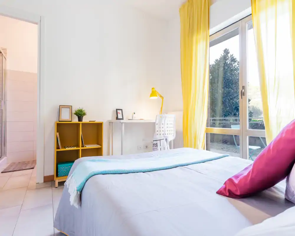 Bright, neatly staged bedroom with large window and yellow curtains; comfortable bed with colorful cushions and a small desk by the window ideal for remote work.