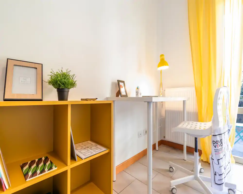 Bright, tidy home-office corner in a living area with a white desk, yellow lamp and matching shelving. Natural light and warm yellow accents create an inviting workspace.