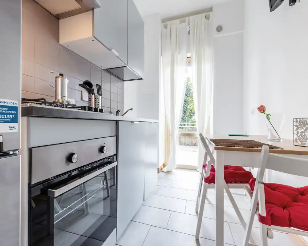 Bright modern kitchen with built-in oven, countertop and a small dining area with red-cushioned chairs; natural light from a doorway creates a welcoming atmosphere.