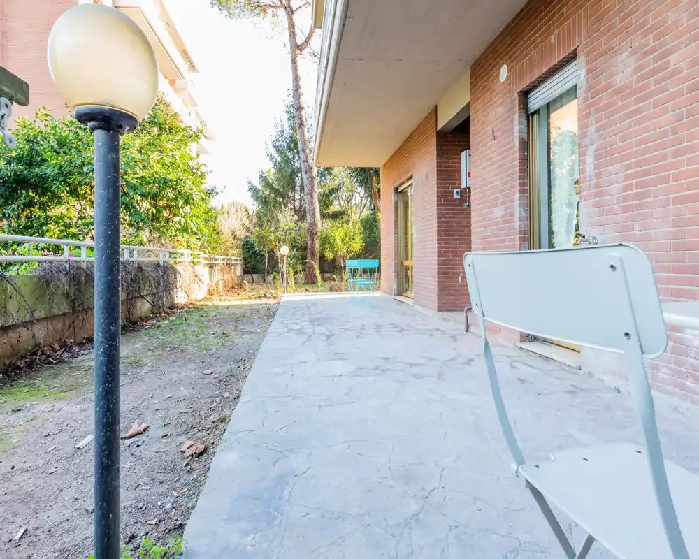 Sunny ground-floor terrace with stone paving, a white folding chair in the foreground and a small garden area with trees and shrubs — ideal for relaxing outdoors.