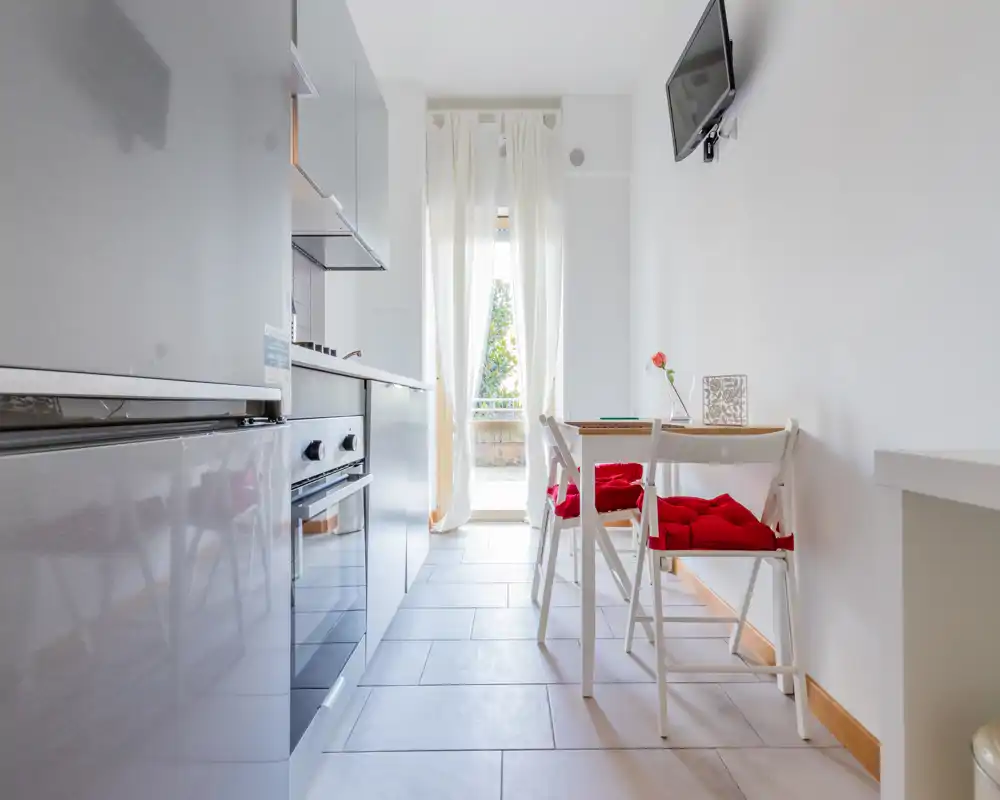 Bright modern kitchenette with integrated appliances and a small dining corner featuring white folding chairs with red cushions and a door letting in natural light.