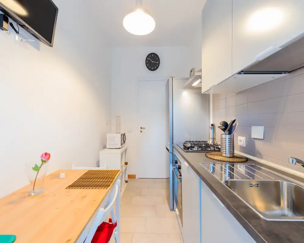 Bright compact kitchen with a stainless-steel sink, gas hob and a wooden dining counter for two; modern white cabinets and neat styling create a clean, functional space.