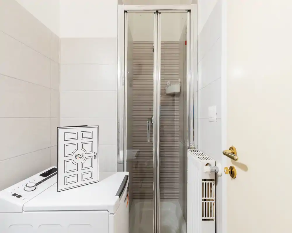 Compact bathroom with a glass shower cabin, white tiles and a washing machine; bright, clean and well-lit space ideal for rental listings.