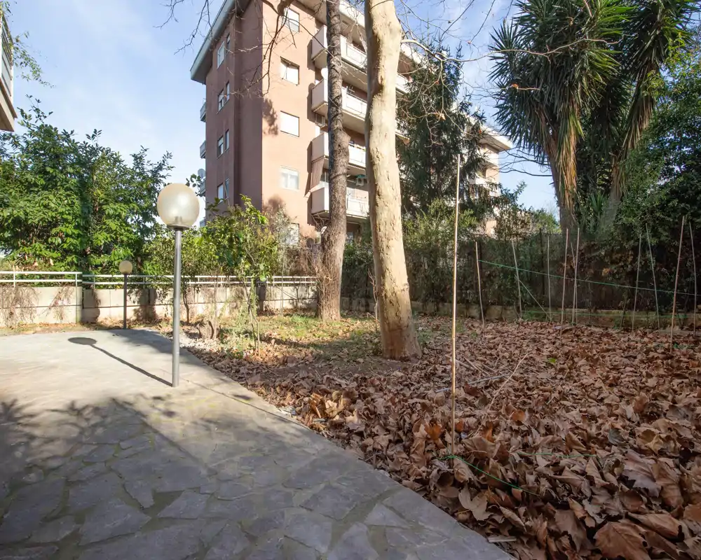 Sunny private garden area with paved patio, mature trees and leaf-covered planting beds — suitable for outdoor relaxation or light gardening.