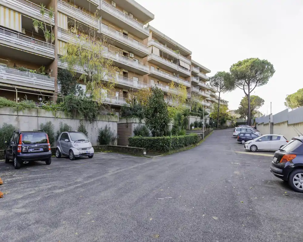 Sunny residential parking area in front of an apartment building, with marked parking bays, hedges and several parked cars — suitable to show available outdoor parking.