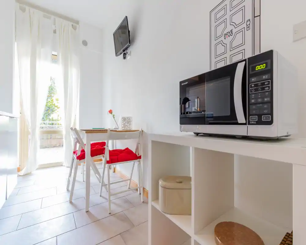 Compact, bright kitchenette with a small dining table, microwave on shelving, and natural light from a balcony door — ideal for short stays.