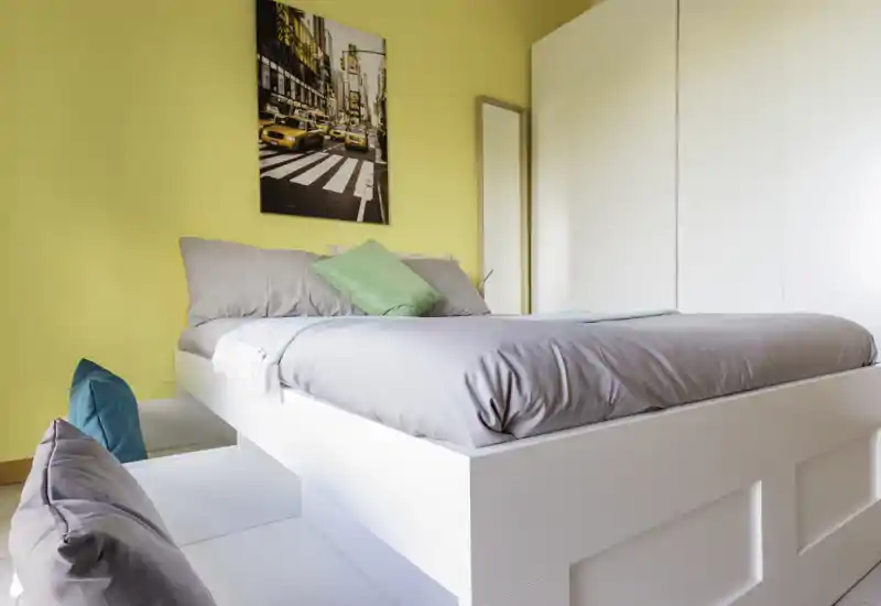 Bright, modern bedroom with a raised white bed frame, grey bedding and colorful accent cushions against a warm yellow wall. The room features a large white wardrobe and a decorative city print, creating a fresh, inviting atmosphere.