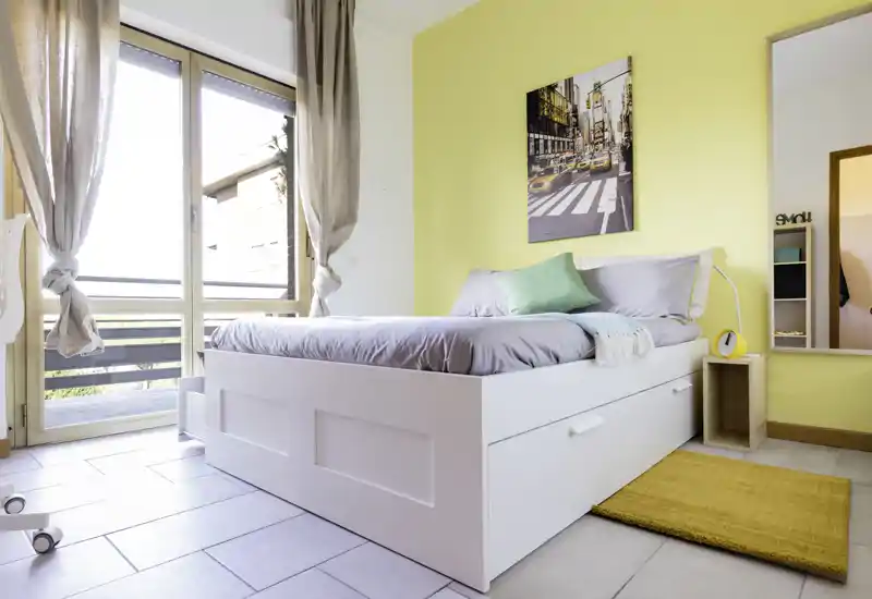 Bright, modern bedroom with a white storage bed, soft gray bedding and a yellow accent wall. Large window with sheer curtains floods the room with natural light; cozy rug and cushions complete the look.