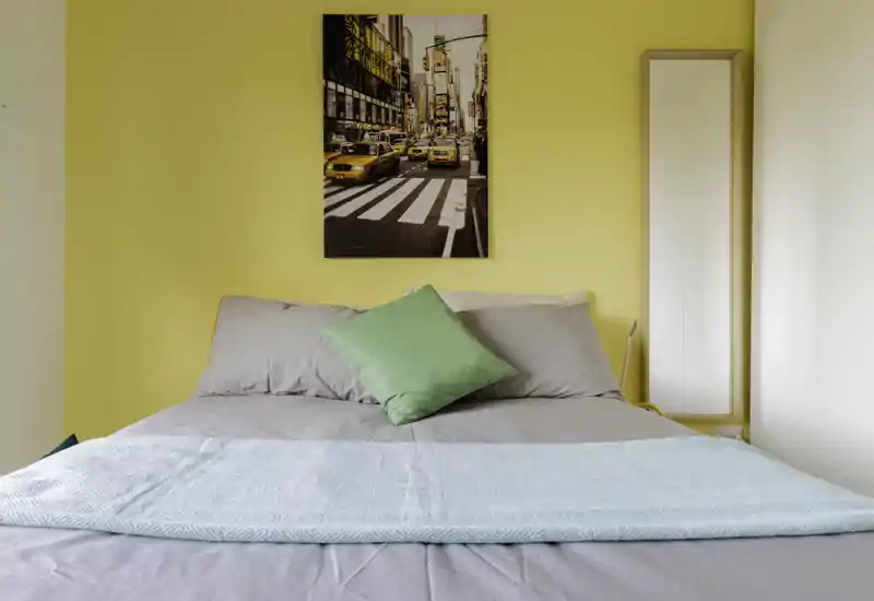 Bright, neatly made bedroom with a soft grey bed, pastel throw and a decorative green cushion against a warm yellow wall; framed city artwork and a full-length mirror complete the composition.